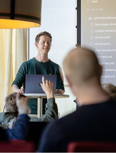 Föreläsare presenterar inför publik med kod på stor skärm.