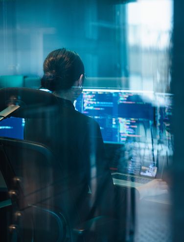 Person i kontorsstol tittar på datorskärmar med kod, sedd genom glas.