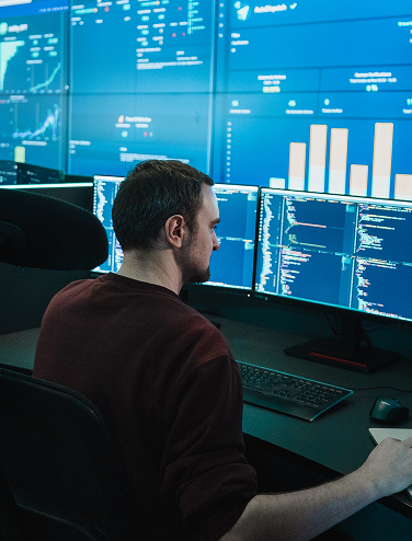Person arbetar vid flera bildskärmar med kod och datadiagram i ett modernt kontor.